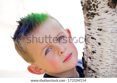 Young boy with blue and green spiky hair hugging birch tree trunk