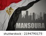 abstract silhouette of the city with text Mansoura near waving colorful national flag of egypt on a gray background. 3D illustration