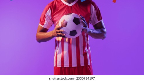 African American man holds a soccer ball, with copy space. Athlete in a red and white jersey stands against a purple background. - Powered by Shutterstock