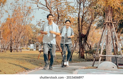 Asian Housewife Rides Electronic Scooter Behind Her Jogging Run Husband In Colorful Foliage Autumn Garden. Watercolor Texture Art Of Cheerful Middle Aged Couple Family Happy Exercising In Autumn Park.