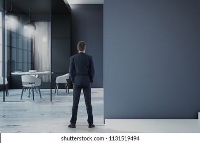 Back View On Businessman Standing On Wooden Floor In Modern Office Near Blank Dark Wall. 