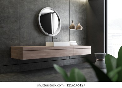 Bathroom corner with concrete walls and floor and white angular sink standing on wooden countertop with round mirror above it. 3d rendering