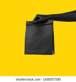 Black Hand Holding Paper Bag For Takeaway Isolated On Yellow Background. Food Delivery Service Concept Banner. 3d Illustration.