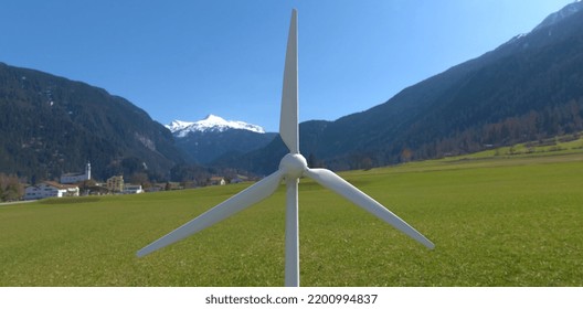 The Blades Of An Industrial Wind Turbine Against The Backdrop Of A Mountain Village. Renewable Energy Plus Self-contained Power Supply. 3d Render.