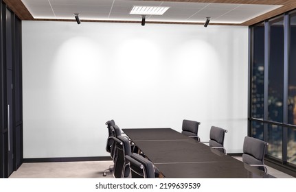 Blank White Wall Mockup In Dark Modern Office With Windows And Bright Spotlights. Empty Company Meeting Room 3D Rendering