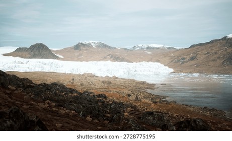 A breathtaking view of a massive glacier flows into a tranquil body of water. Surrounding mountains display earthy tones, creating a beautiful contrast. Clouds softly drift in a clear sky.