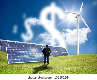 Businessman Looking At Dollar Signs Made Of Clouds In A Field With Solar Panels And Wind Turbine