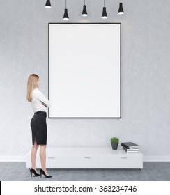 Businesswoman Standing In Front Of A Big Blank Frame Hanging On The Wall Above A Shelf, Five Lamps On The Ceiling. Concrete Background. Front View. Concept Of Watching.