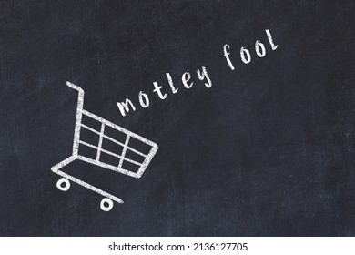 Chalk Drawing Of Shopping Cart And Word Motley Fool On Black Chalboard. Concept Of Globalization And Mass Consuming.