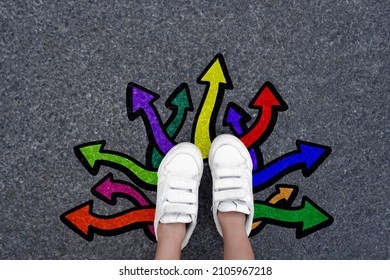 Choice Concept. Feet Walking Away And Colorful Graffiti Direction Arrows On Road Background. Selfie Children Shoes Top View. Journey Beginning And Future New Way.