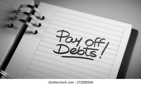 Closeup of a personal agenda setting an important date representing a time schedule. The words Pay off debts written on a white notebook to remind you an important appointment.