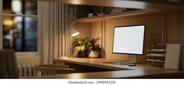 A computer with a white screen mockup and accessories on a wooden table, illuminated by a table lamp in a home office workspace at night. 3d render, 3d illustration