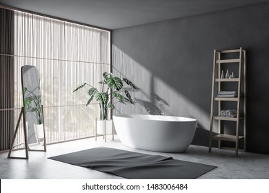 Corner of minimalistic bathroom with gray walls, concrete floor, light wooden blinds, comfortable white bathtub, shelves with towels and vertical mirror. 3d rendering