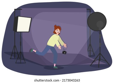 Cute Awkward Girl With Red Hair In Glasses Looks Frightened, Tries To Catch A Falling Camera. Dark Photo Studio With Lighting. Minimalist Style
