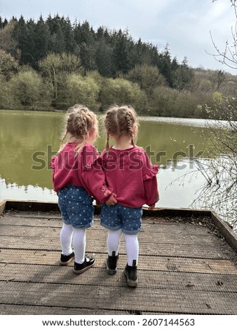 Description:
Two young children stand side by side on a wooden platform, looking out over a still lake surrounded by trees. Their matching outfits and close stance create a sweet, peaceful moment.

