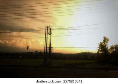 Dramatic sunset view in the countryside, featuring electric poles with wires crossing the golden sky, silhouettes of palm trees, and a stretch of green rice fields. The combination of orange and yello