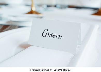 An elegantly arranged wedding reception table showcases a sophisticated white place card with 'Groom' beautifully scripted, surrounded by upscale dining ware and floral decorations. - Powered by Shutterstock