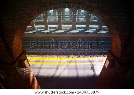 An evocative composition highlights the contrasting textures and forms as sunlight streams through an arched window, offering a voyeuristic view of a busy railway platform below filled with commuters.