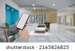 A female hand holding a smart mobile phone or cellphone white screen mockup over blurred modern clinic or health care centre reception lobby in the background. 3d rendering, 3d illustration