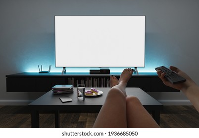 First Person Shot Of A Girl Sitting In An Armchair Watching TV, With Her Legs Resting On The Table And Pointing The Remote Control. Living Room With Modern Lighting. Mockup. 3d Rendering