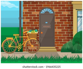 Front Door To A Brick House, Porch With Steps And A Yellow Bike Standing Next To It. Bushes To The Right, Grass In The Foreground. In The Background Is The Sky