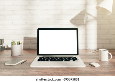 Front view of wooden hipster office desk top with blank white laptop display, coffee cup, supplies and other items. Brick wall background with shadows and sunlight. Mock up, 3D Rendering 