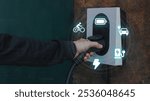 Hand plugs electric car charger as icons for cars and bikes display above, green metal background with subtle texture adds depth, vehicle charging, eco mobility, tech innovation