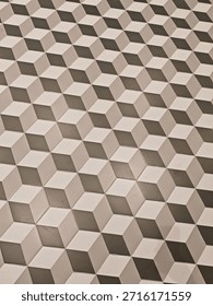 A high-angle, full-frame shot of a tiled surface featuring a tumbling blocks or reversible cube pattern. The design uses three distinct shades—white, light grey, and dark charcoal grey—to create a convincing 3D isometric effect. The repeating rhombic shapes give the viewer the impression of looking down at a series of stacked cubes or an architectural structure. The lighting is soft and warm, highlighting the matte texture of the tiles and the thin grout lines connecting them.