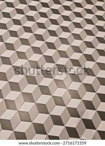 A high-angle, full-frame shot of a tiled surface featuring a tumbling blocks or reversible cube pattern. The design uses three distinct shades—white, light grey, and dark charcoal grey—to create a convincing 3D isometric effect. The repeating rhombic shapes give the viewer the impression of looking down at a series of stacked cubes or an architectural structure. The lighting is soft and warm, highlighting the matte texture of the tiles and the thin grout lines connecting them.