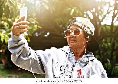 ILLUSTRATION Portrait Senior Asian Woman Selfie. Thai old female wearing trendy hoodie jacket,cap,sunglasses
and holding smartphone take photo in green nature garden park. Asia Grandmother happy smile - Powered by Shutterstock