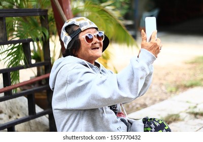 ILLUSTRATION Portrait Senior Asian Woman Selfie. Thai Old Female Wearing Trendy Hoodie Jacket,cap,sunglasses
And Holding Smartphone Take Photo In Green Nature Garden Park. Asia Grandmother Happy Smile