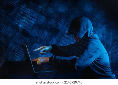 Image Of A Man In A Black Hoodie Pointing To The Screen Of A Laptop Computer At Home And Remotely Controlling It, Under A Blue Glowing Binary Data In A Glass Cube On Chains.