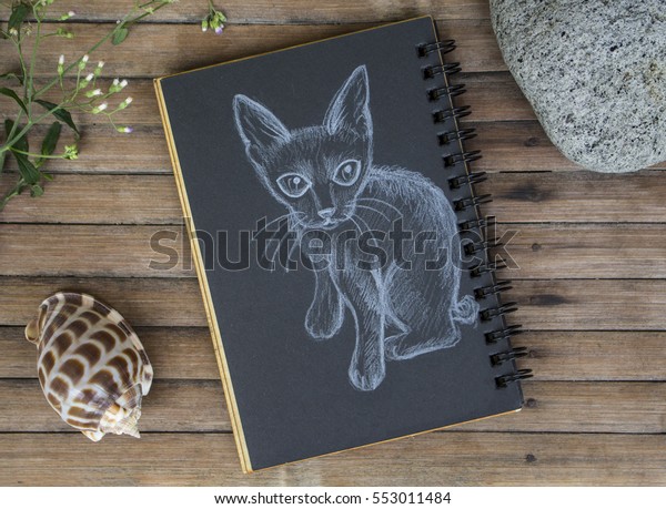 Kitten with small tail hand-drawn illustration. Cat\
by white chalk on black paper. Black paper notepad on wooden\
background. Vintage wooden table with artwork. Cute cat drawing.\
Table of artist\
photo
