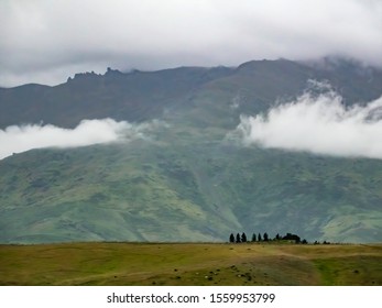 Land Of Mystery: Green Valley With Evergreen Windbreak Hiding Farm Buildings, Maybe, Near Misty Mountainside Under Rain Clouds In The Otago Region Of New Zealand, With Light Digital Painting Effect