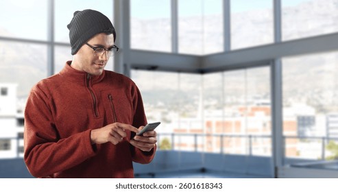 Man in beanie using smartphone in modern office with large windows. Technology, business, workplace, communication, contemporary, professional