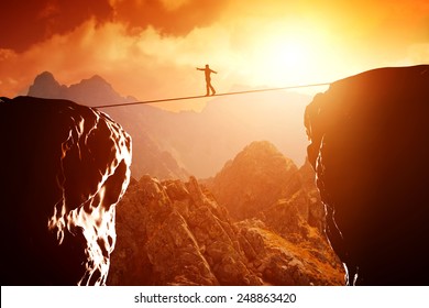 Man Walking And Balancing On Rope Over Precipice In Mountains At Sunset. Concept Of Business, Risk Taking, Challenge, Concentration.