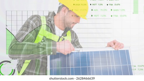 Man wearing yellow hard hat, safety vest adjusting solar panel in studio, with financial charts. Industrial, sustainability, technology, innovation, analytics, engineering, environment