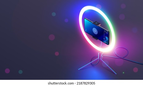 Phone On RGB Led Ring Light On Tripod On Table. Smartphone Streams Video In Internet. Vlogging Concept. Color Smart Lamp For Shoot A Video Or Selfie Through Camera Phone In The Dark On Desk.