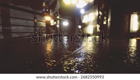 Raindrops create a glossy surface on the street, mirroring the vibrant city lights. A person walks by, adding life to the serene nighttime atmosphere filled with reflections.