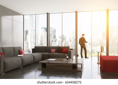 Rear View Of A Businessman Standing Near A Panoramic Living Room Window And Looking At His Smartphone Screen. There Are Two Gray Sofas And A Large Coffee Table. 3d Rendering, Mock Up, Toned Image