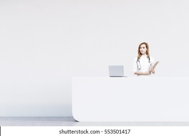 Red Haired Woman Receptionist Standing At A Counter In A Hospital. There Is A White Wall Behind Her And A Laptop On The Desk. 3d Rendering. Mock Up