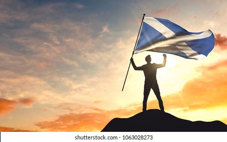 Scotland Flag Being Waved By A Man Celebrating Success At The Top Of A Mountain. 3D Rendering