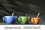 Set of three colored tea mugs with spoons inside stand in a row on a dark background.