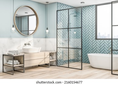 Side view on eco interior design of bathroom with wooden frame of round mirror on blue wall, wooden sink cabinet and floor, white bath and herringbone tiles wall behind glass partitions. 3D rendering - Powered by Shutterstock