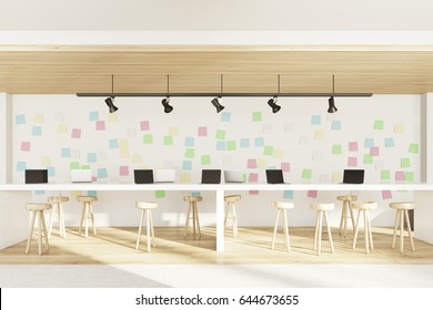 Side view of an open office interior with two bar tables with stools, laptops standing on them, a light wooden floor and a wall with sticky notes. 3d rendering