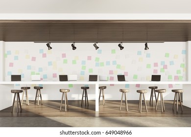 Side view of an open office interior with two bar tables with stools, laptops standing on them, a dark wooden floor and a wall with sticky notes. 3d rendering