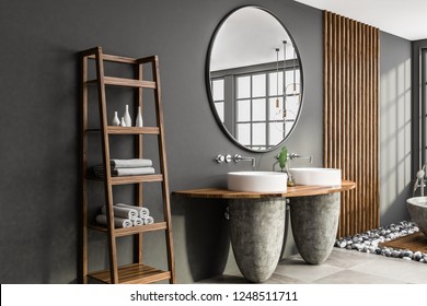 Side view of white and stone double bathroom sink standing on wooden shelf in room with gray walls and large round mirror. Shelves with towels. 3d rendering