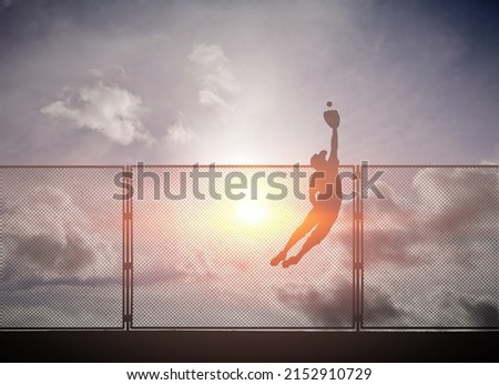 Silhouette of a baseball player diving to catch a ball from going over the outfield fence against a surreal sunset sky.