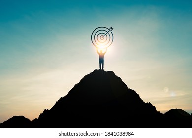 Silhouette of businessman holding target board on the top of mountain with over blue sky and sunlight. It is symbol of leadership successful achievement with goal and objective target.