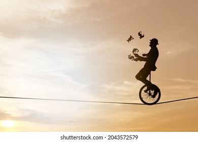 Silhouette Of A Businessman In Suit And Bowler Hat Juggling Currency Symbols While Riding A Unicycle On A Tightrope Against A Dreamy Sunset Sky For The Concept Of Uncertain Risky Business World.